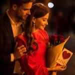 handsome man hugging happy girlfriend holding bouquet of roses and card with love inscription and heart symbol